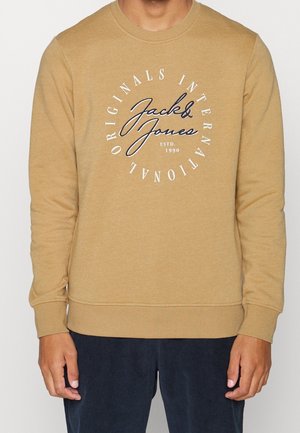 Beiger langärmliger Pullover mit dem weißen und schwarzen Schriftzug „Jack & Jones Originals International Estd. 1990“, getragen zu dunkler Hose.