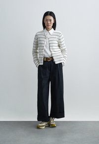 Witte cardigan met horizontale strepen, dichtgeknoopt, gedragen over een wit overhemd met kraag. Losvallende donkergekleurde broek en beige sneakers met gele accenten.