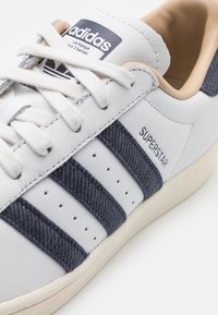 Sneaker in pelle bianca con accenti in denim blu e tre strisce distintive. Presenta una superficie testurizzata, perforazioni per la traspirazione e un'etichetta con logo.