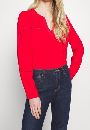 Rotes langärmliges Oberteil mit V-Ausschnitt, das an den Schultern mit strukturiertem Rippmuster versehen ist, kombiniert mit dunkelblauen Denim-Jeans.