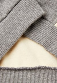 Pullover grigio con collo e polsini a coste. Superficie morbida e testurizzata combinata con una fodera interna liscia e cremosa. Cuciture visibili lungo i bordi.