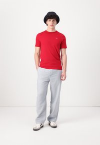 Camiseta roja de manga corta, corte relajado, combinada con pantalones gris claro y zapatillas blancas con acentos negros. El modelo lleva puesto un sombrero de cubo negro.