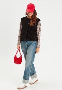 Gilet di pile nero con bottoni frontali, abbinato a una camicia a maniche lunghe con stampa leopardata, jeans blu e sneakers bianche. Accento di una borsa rossa.