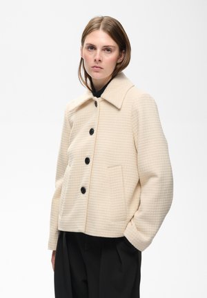 Jeune femme portant une veste crème texturée avec des boutons noirs et un pantalon noir, debout avec les mains dans les poches devant un fond blanc uni.