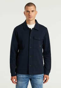 Marineblauer Baumwolljacke mit durchgehender Knopfleiste, zwei vorderen Taschen, Kentkragen und gerippten Bündchen, getragen über einem weißen Hemd und blauen Jeans.