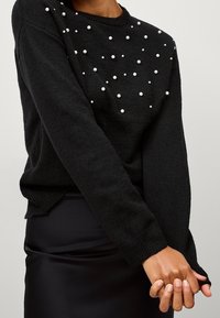 Schwarzer Pullover mit langen Ärmeln, der ein verstreutes Design aus weißen Perlenakzenten auf der Vorderseite aufweist. Strukturierter Strickstoff mit gerippten Bündchen.