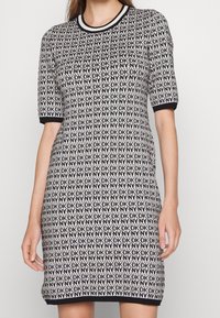 Femme portant une robe en maille noire et blanche à manches courtes avec un motif répété de logo "DKNY" et des bordures noires aux manches et à l'ourlet.
