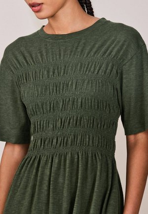 Femeie purtând o rochie verde închis cu mâneci scurte și un corset texturat cu pliuri adunate.