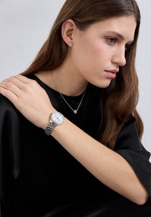 Joven mujer que lleva un reloj de plata, un collar de plata con colgante y pequeños pendientes de botón, vestida con blusa de satén negra, mirando hacia un lado.