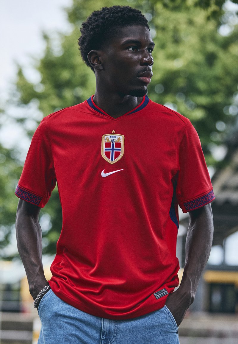 Maglia sportiva rossa con scollatura a V, caratterizzata da accenti blu e un motivo testurizzato sulle maniche. Presenta uno stemma e il logo Nike.