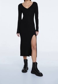 Schwarzes, geripptes Langarmkleid mit tiefem V-Ausschnitt und seitlichem Schlitz. Kombiniert mit schwarzen schnürbaren Kampfstiefeln. Glatte Textur, figurbetonte Silhouette.