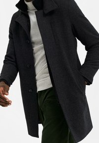 Tmavě šedý kabát z texturované látky, s knoflíkovou přední částí, límcem a bočními kapsami. Nošený přes světle šedý svetr a zelené kalhoty.