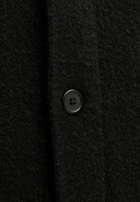 Schwarzer Wollmantel mit strukturierter Oberfläche, verziert mit einem runden schwarzen Knopf mit vier Löchern, der die fuzzy Optik des Stoffes zur Geltung bringt.