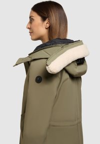 Oliven grøn parka med en blød hvid fleeceforet hætte, sort knap og lynlås foran. Har en glat tekstur og sidelommer.