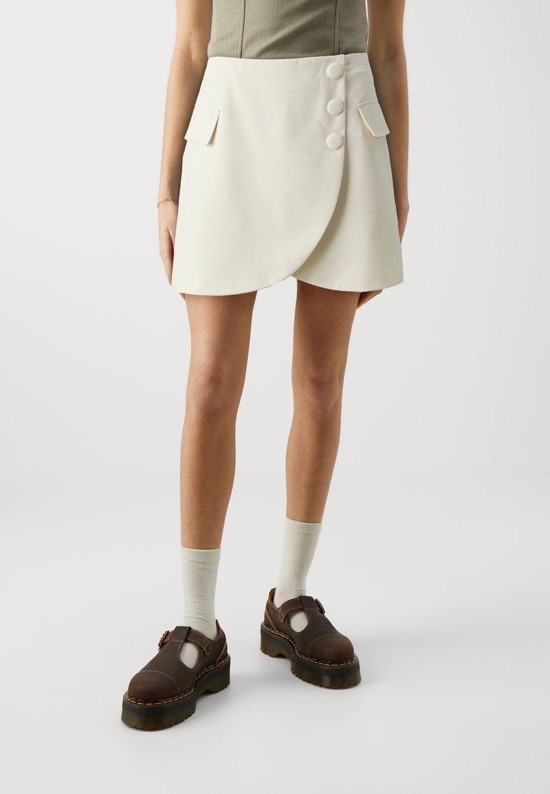 Femme portant une jupe portefeuille crème avec trois boutons et des poches à rabat, assortie à des chaussettes blanches et des chaussures à plateforme marron foncé.