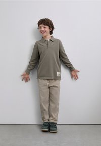 Tricou polo cu mâneci lungi, de culoare măslinie, pantaloni bej și pantofi verzi. Tricoul are guler și o etichetă mică pe tiv.