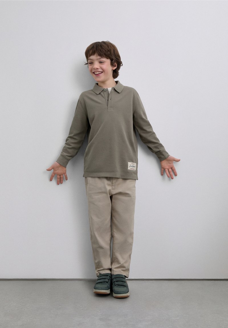 Tricou polo cu mâneci lungi, de culoare măslinie, pantaloni bej și pantofi verzi. Tricoul are guler și o etichetă mică pe tiv.