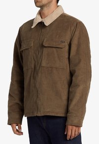 Chaqueta de pana marrón con cuello de piel sintética beige, dos bolsillos en el pecho, cierre de cremallera y textura acanalada. Diseño informal para el clima fresco.