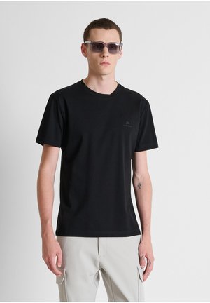 Joven con gafas de sol oscuras, camiseta negra de manga corta y pantalones cargo de color claro, de pie frente a un fondo liso.