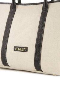 Kremowa torba tote z płótna z ciemnymi skórzanymi uchwytami i akcentami. Posiada złote logo "VENEZIA" na prostokątnym czarnym patchu.