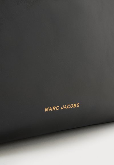 Surface en cuir noir avec petit texte doré "MARC JACOBS" embossé près du bord inférieur.