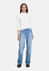 Weißer gerippter Rollkragenpullover kombiniert mit hellblauen geraden Jeans und braunen Plateauschuhen, stehend vor einem schlichten Hintergrund.