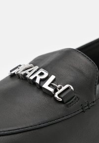 Chaussure noire en cuir à enfiler, présentant un emblème chromé brillant "KARLO" comme détail marquant, avec des coutures cousues et une surface texturée.