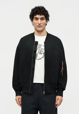 Mężczyzna z kręconymi włosami, ubrany w czarną kurtkę na zamek błyskawiczny na białą koszulkę z grafiką i czarne spodnie, stojący na jednolitym białym tle.