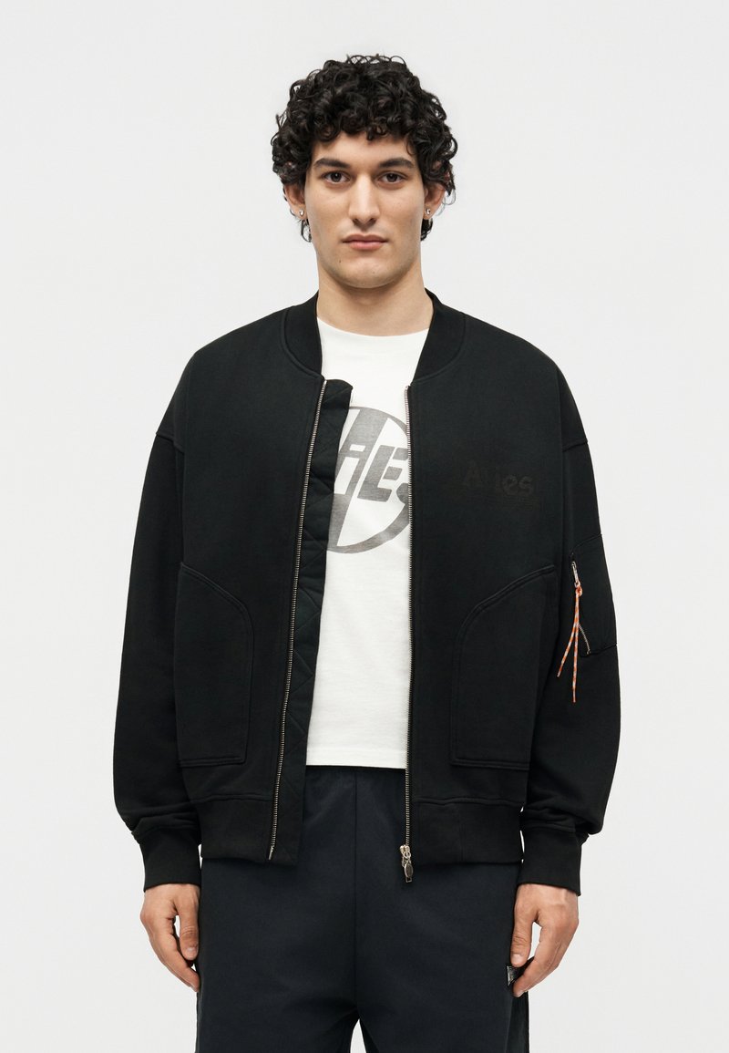 Hombre con cabello rizado que lleva una chaqueta negra con cremallera sobre una camiseta blanca con gráfico y pantalones negros, de pie frente a un fondo blanco liso.