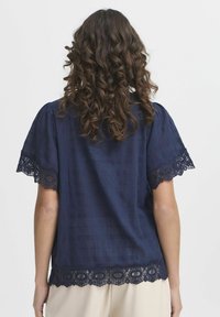 Blusa blu navy con maniche corte, caratterizzata da una finitura in pizzo intorno alle maniche e alla vita, realizzata in tessuto testurizzato con una finitura decorata.