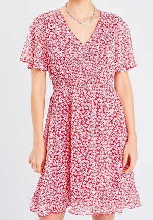 Femme portant une robe rouge à motifs floraux blancs, manches courtes flottantes, un décolleté en V, et un collier chaîne argent.
