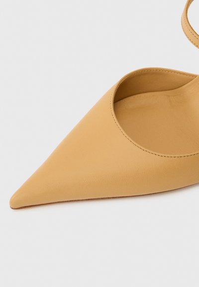Beige kožené baleríny s špičatou špičkou a nízkým výstřihem na bílém pozadí, ukazující vnitřek a okraj kotníkového pásku.