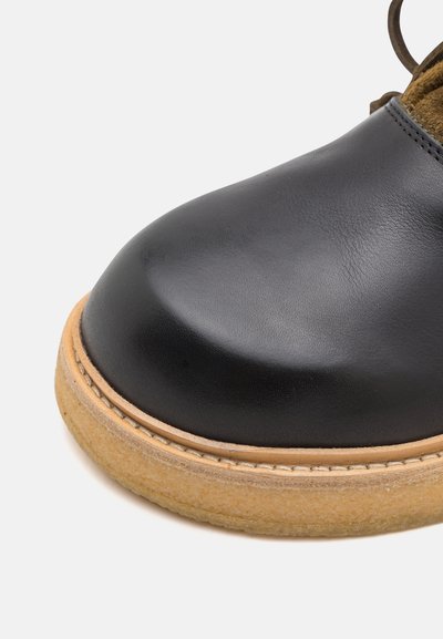 Schwarzer Lederschuh mit abgerundetem Zehenbereich, ausgestattet mit einer hellbraunen Gummisohle und einer Steppnaht am Rand. Weiches Innenfutter ist sichtbar.