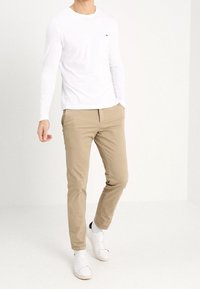 Bílá košile s dlouhým rukávem, béžové kalhoty na míru a bílé tenisky. Na košili je malé logo na hrudi. Hladký textura látky.