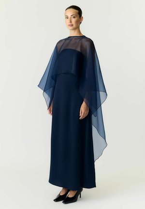 Vrouw staat in een vloerlang marineblauw jurkje met een doorschijnende marineblauwe cape en zwarte pumps tegen een effen achtergrond.