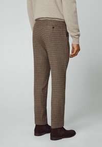 Muž oblečený v kostkovaných hnědo-béžových kalhotách, béžovém svetru a tmavě hnědých semišových chelsea botách, stojící proti jednoduchému světlému pozadí.