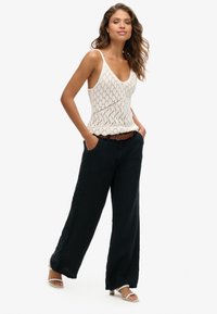 Witte gebreide tanktop met een diamanten patroon, gecombineerd met losse zwarte broek. Model draagt een bruine gevlochten riem en witte hakken.
