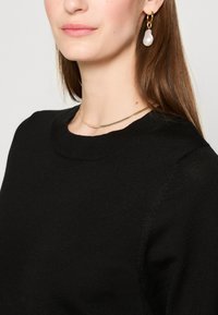 Pulover negru din tricot cu guler rotund, cusături detaliate și o textură netedă. Însoțit de un colier din lanț de culoare aurie și cercei cu perle.