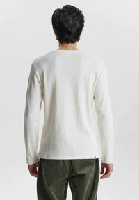 Langärmliges Shirt in Creme mit gerippter Textur, Rundhalsausschnitt und geradem Saum. Kombiniert mit dunkelgrünen Baumwollcord-Hosen.