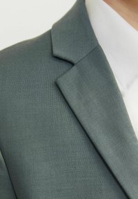 Veste de costume vert foncé en tissu texturé, avec un revers cranté et une chemise blanche partiellement visible en dessous.