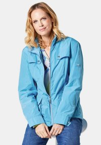Leichte blaue Jacke mit Reißverschluss, Druckknopftaschen und verstellbaren Bündchen. Sie verfügt über einen Stehkragen und eine glatte, glänzende Textur.