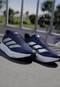 Scarpe sportive blu navy con tre strisce argentate, tomaia strutturata e suola intermedia bianca. Design leggero adatto per la corsa o l'allenamento.