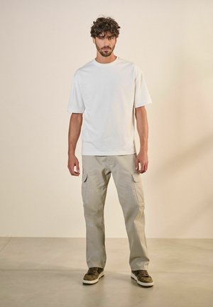 Weißes Baumwoll-T-Shirt mit kurzen Ärmeln, kombiniert mit hellbeigen Cargo-Hosen mit Seitentaschen, getragen mit braunen und weißen Sneakern.