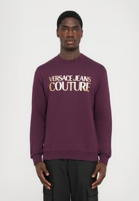 Felpa viola con la scritta dorata "VERSACE JEANS COUTURE", colletto a giro, polsini e orlo a coste, e una finitura morbida e texturizzata.