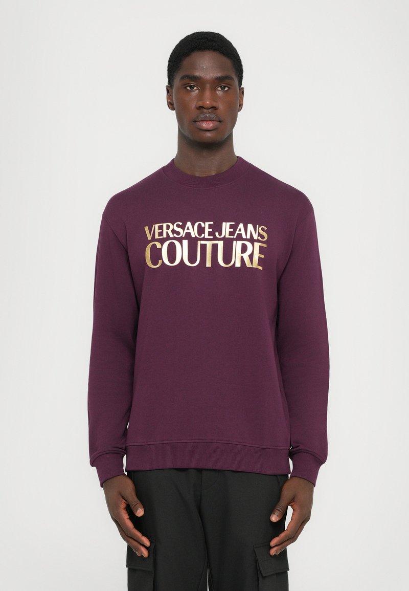Felpa viola con la scritta dorata "VERSACE JEANS COUTURE", colletto a giro, polsini e orlo a coste, e una finitura morbida e texturizzata.