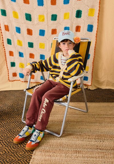 Niño pequeño con suéter de rayas y gorra adornada con flores sentado en una silla plegable frente a un fondo de manta de ganchillo colorida.