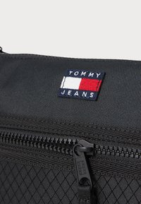 Sac en tissu noir avec une surface texturée, une fermeture éclair et un patch logo TOMMY JEANS bien visible en rouge, blanc et bleu.