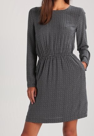 Vrouw draagt een zwarte jurk met lange mouwen, voorzien van een kleine witte stippenprint, elastische taille en haar hand in de zijzak.