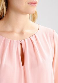 Blouse rose en tissu léger avec un col rond, présentant des plis et une petite ouverture en forme de clé au niveau de la poitrine. Texture lisse.
