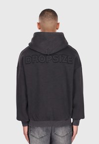 Mand med kort hår iført en mørkegrå hættetrøje med "DROPSIZE" broderet på den øverste del af ryggen og slidte grå jeans.